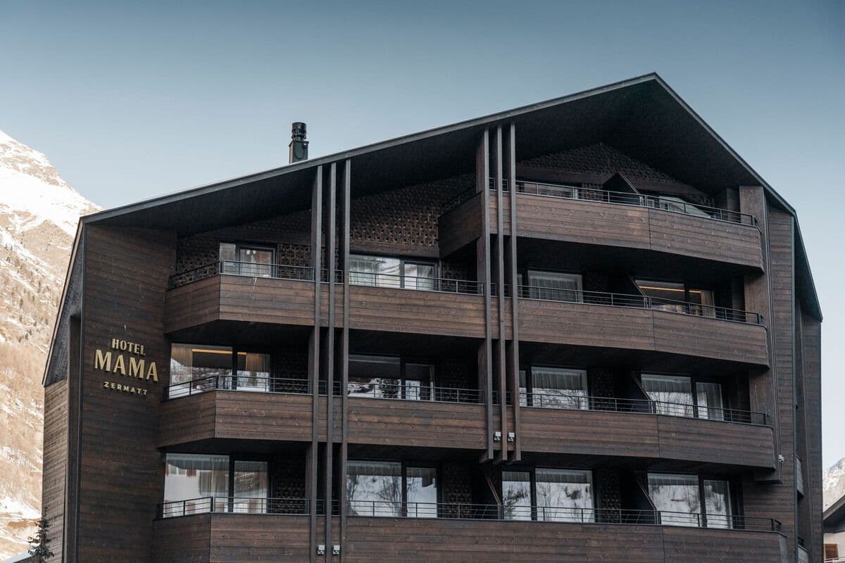 Frontalansicht des Hotel Mama in Zermatt mit dunkler Holzfassade, grossflächigen Balkonen, markanter Dachform und Sicht auf das Hotel Mama Zermatt Logo.