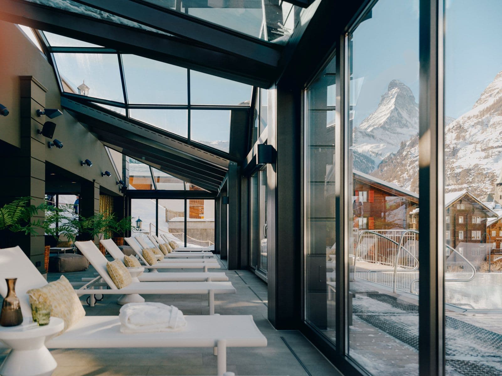 Innenansicht der Entspannungslounge mit weissen Liegestühlen und grossen Glasfenstern, die einen Blick auf das Matterhorn und umliegende Häuser bieten.
