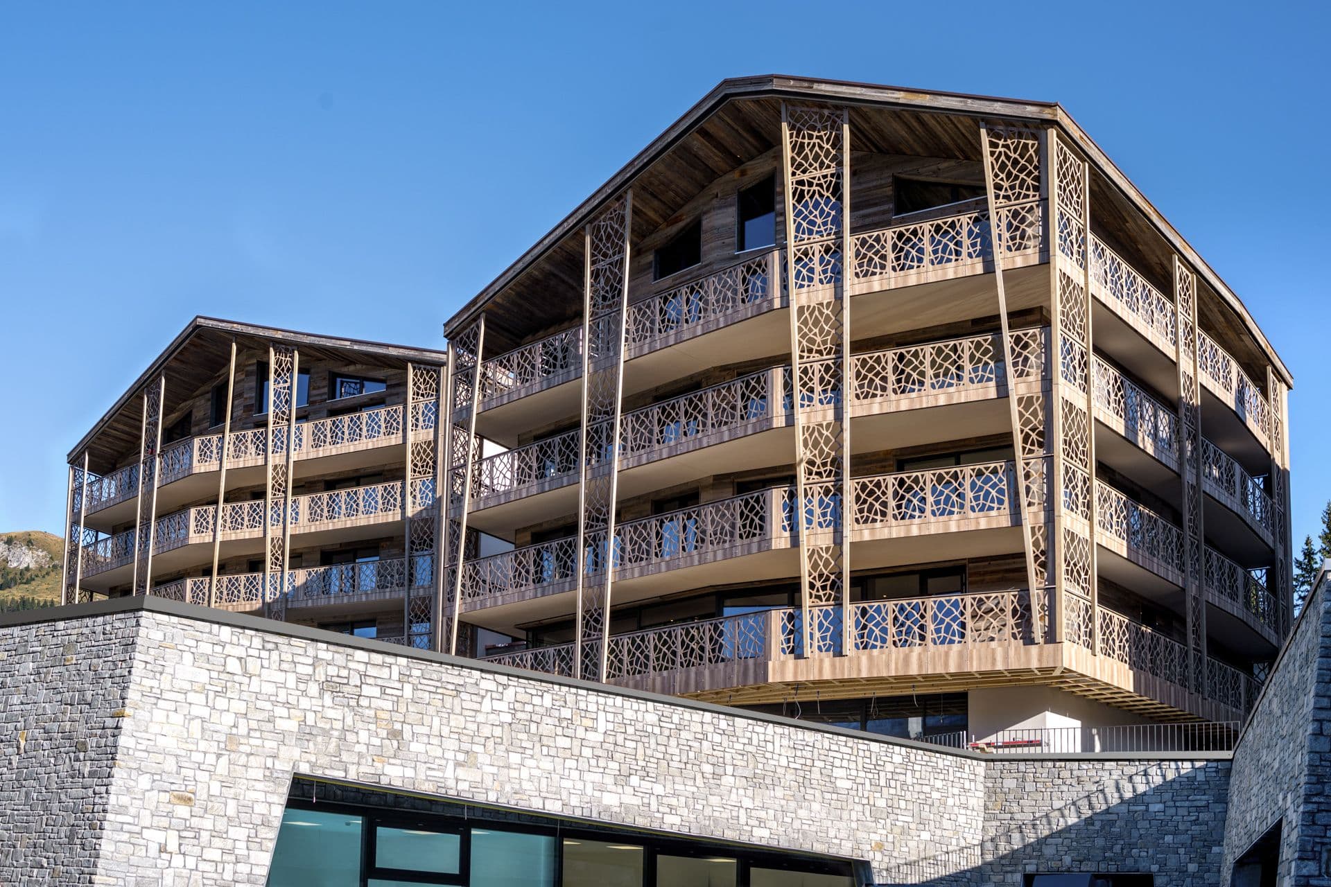 Aussenansicht des Valsana Hotel & Appartements in Arosa mit mehreren Stockwerken und grossen Balkonen. Die Fassade kombiniert eine Holzverkleidung mit dekorativen Metallgittern an den Balkongeländern. Das Gebäude steht auf einem Sockel aus hellen Natursteinmauern.